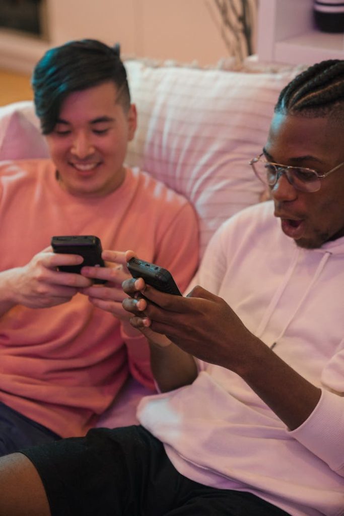 Two friends laughing and playing video games on smartphones while sitting together on a cozy couch indoors.